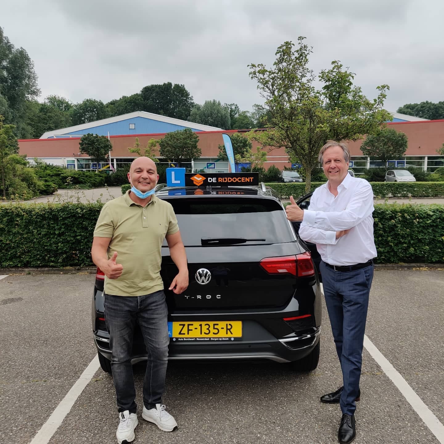Geslaagde leerling en instructeur met duim omhoog bij de lesauto van De Rijdocent